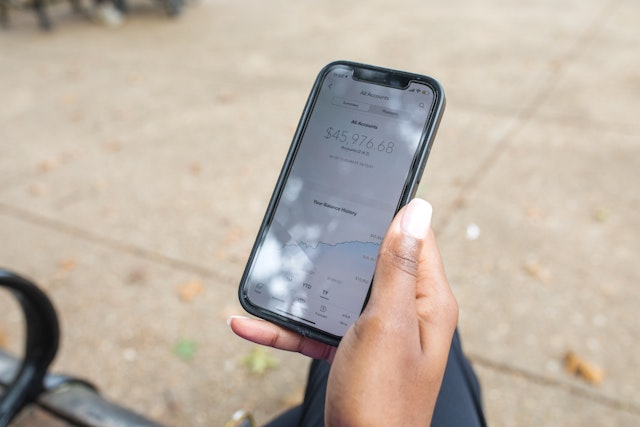 Gifted collabs. Black woman sitting on a bench looking at her phone that displays a financial website. The account balance is $45,976.68.