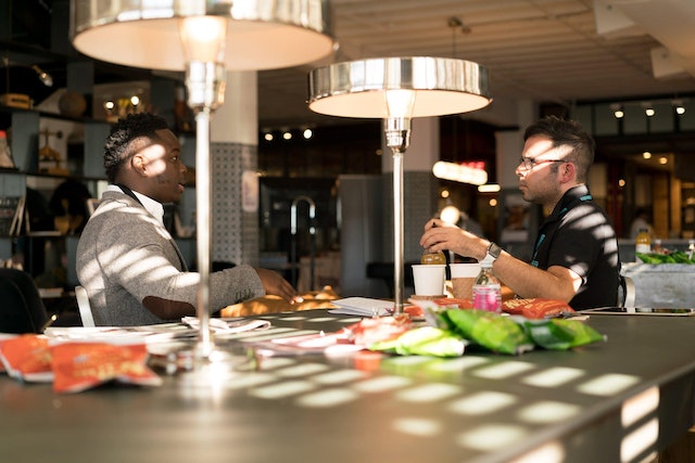 Food industry venture capital. Two people sit at a table having a discussion. There are coffee cups, water bottles and snacks in the background. There are two lamps on the table. 