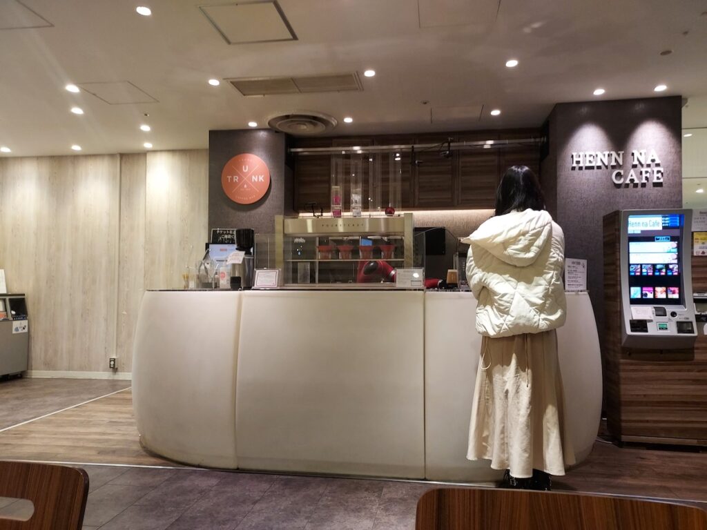 The front of Henn na cafe in Shibuya, Tokyo, Japan. Woman in white coat and beige skirt standing at the bar waiting for her drink to be made by the coffee robot
