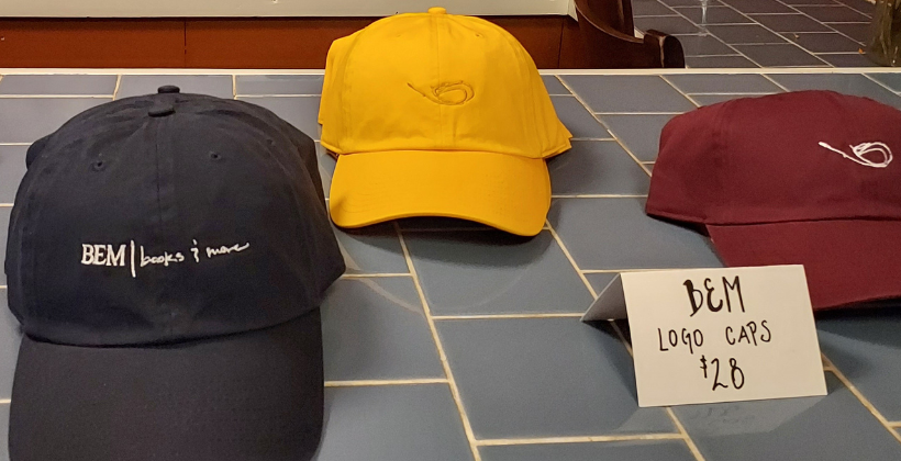 BEM books and more merch. There are three baseball caps, one blue, yellow then red. There's a white paper sign that says, BEM logo caps $28.