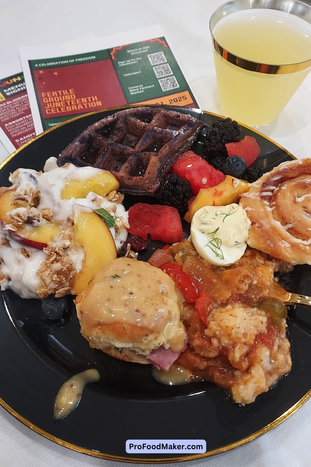 An assortment of brunch food including fresh fruit, a biscuit, pastries and waffles on a black plate outlined in gold. 