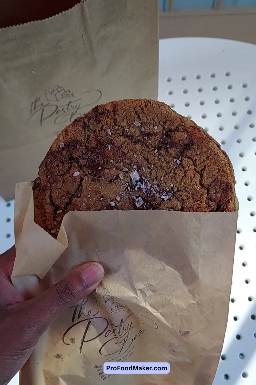 Large chocolate chip cookie with sea salt flakes in a small brown wrapper with The Pastry Box logo stamped.