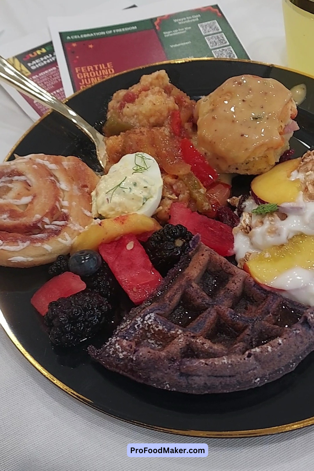 An assortment of brunch food including fresh fruit, a biscuit, pastries and waffles on a black plate outlined in gold. 